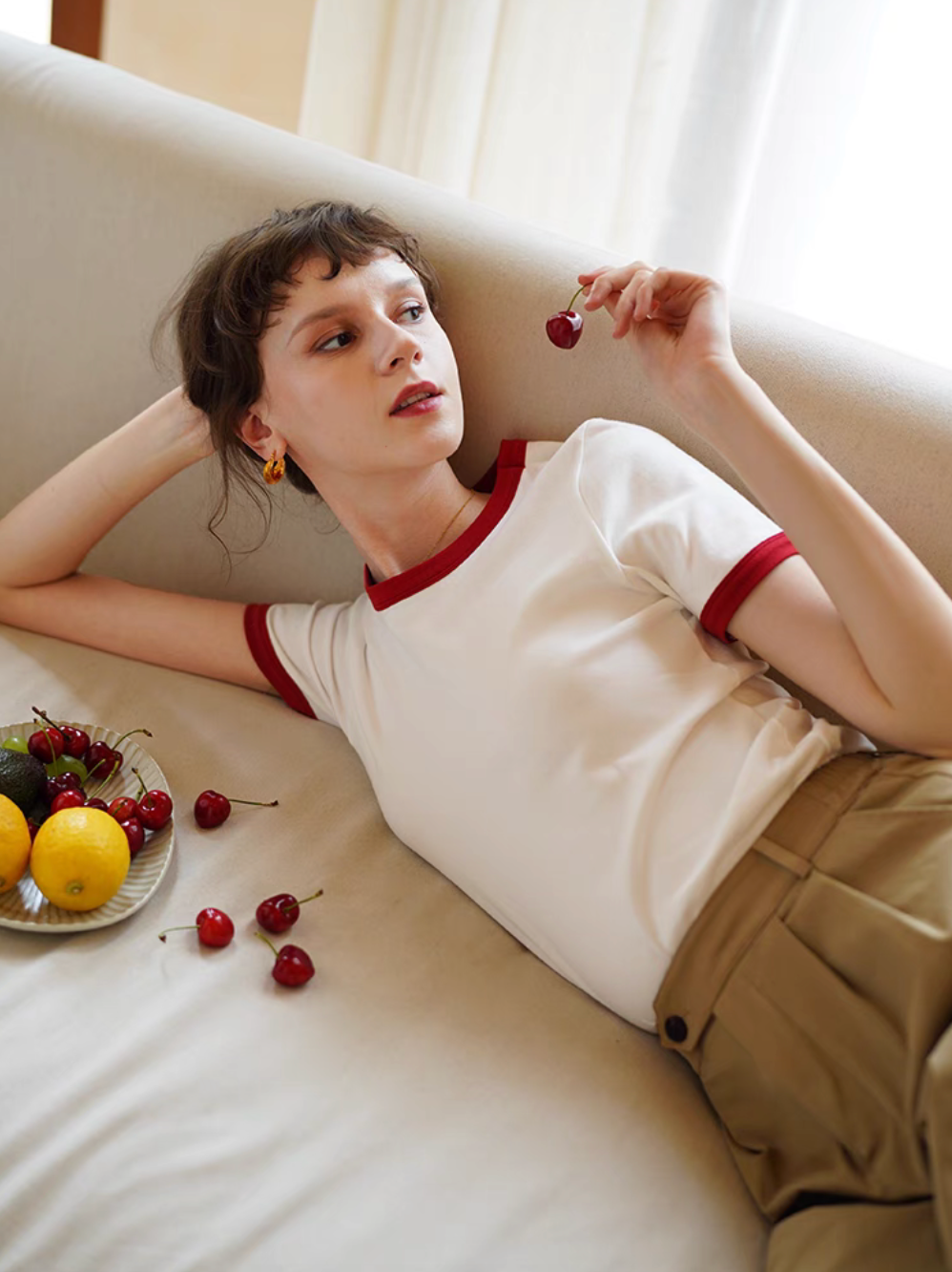 Model reclining on sofa wearing Vintage French Contrast Collar Tee in white with red trim holding cherry with fruit plate creating a relaxed vintage mood