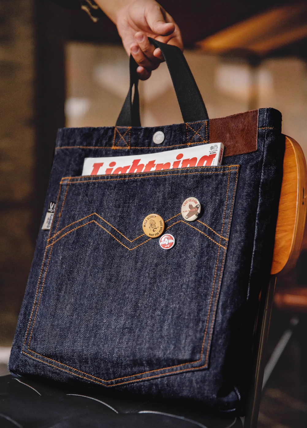 Denim messenger tote bag in denim blue featuring front pocket with decorative pins and magazine inside