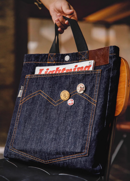 Denim messenger tote bag in denim blue featuring front pocket with decorative pins and magazine inside