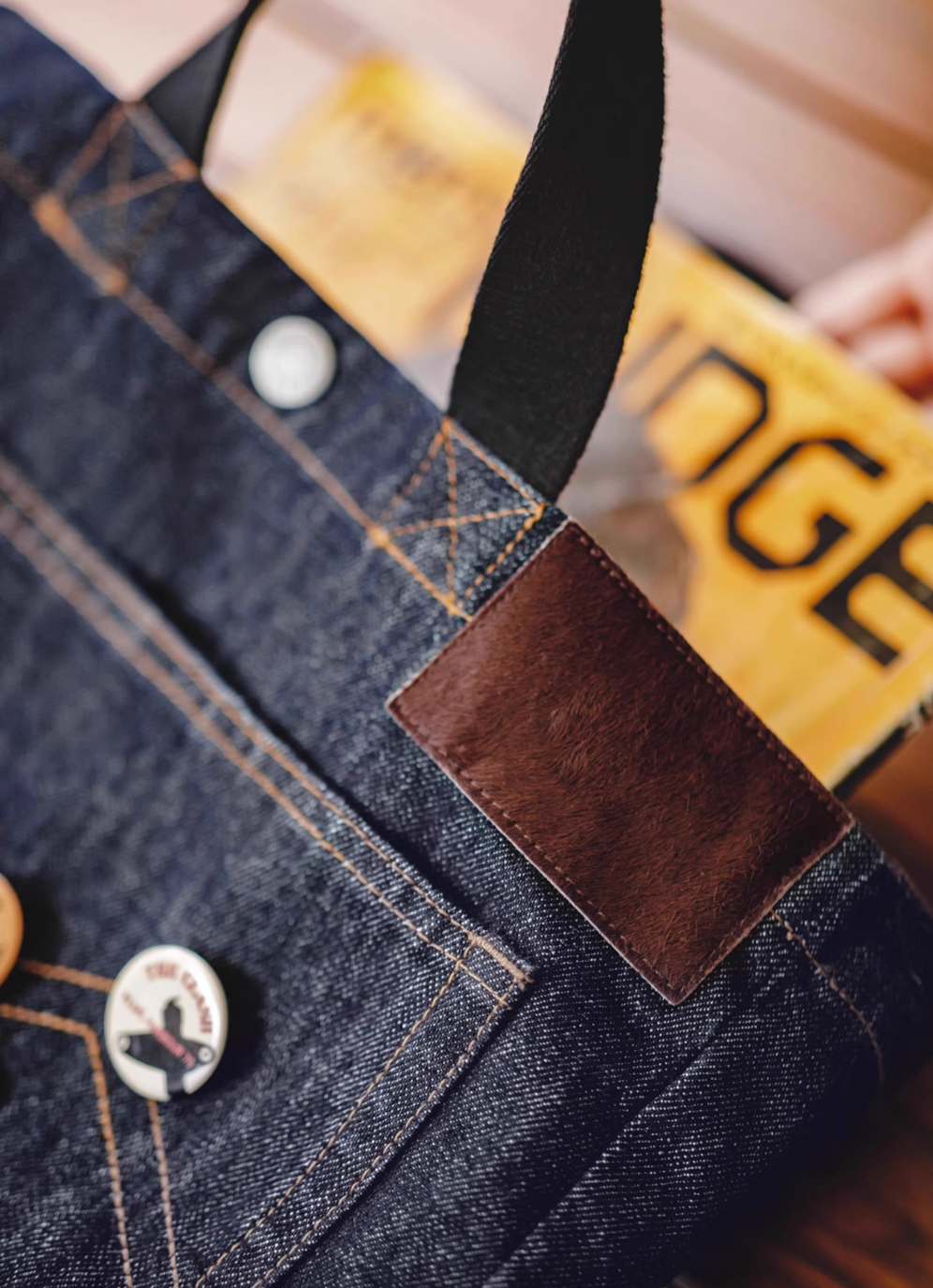 Close-up of denim tote bag showing brown leather patch, stitching detail, and handle attachment