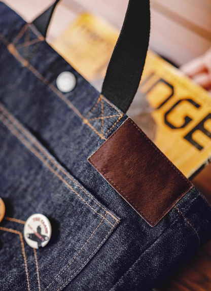 Close-up of denim tote bag showing brown leather patch, stitching detail, and handle attachment