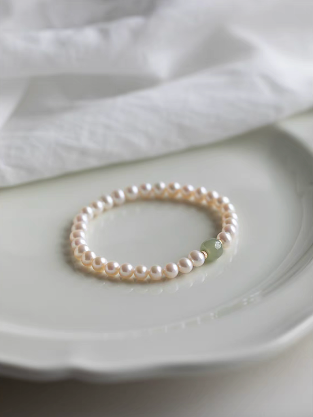 Bracelet displayed on ceramic plate showing natural color pearls with central jade bead in soft lighting.