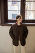Model wearing Vintage Meadow Jacket in dark brown green standing by window, featuring diamond quilted pattern, corduroy collar, and relaxed fit silhouette