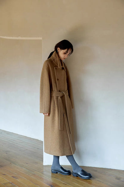Side profile of woman wearing khaki long Willow Lane Wool Coat standing by wall, featuring clean lines, wrap silhouette, and soft structured wool blend