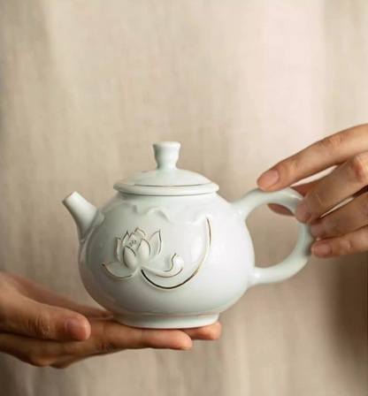 Yingqing Lotus Relief Ceramic Teapot in white color with embossed lotus design and gold detailing, front view held in hands showing rounded body and lid