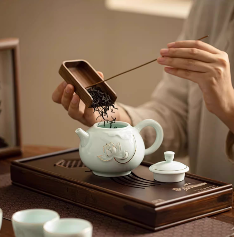 Yingqing Lotus Relief Ceramic Teapot in white with lid removed as loose tea leaves are added using a wooden scoop on a tea tray setup